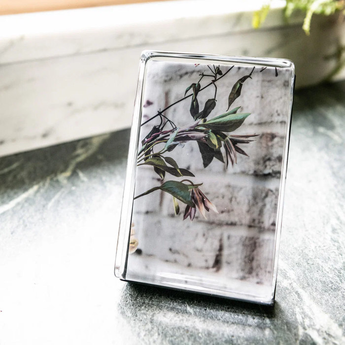 The Simon Pearce Woodbury Photo Block Vertical displays a botanical image of green leaves and flowers, handcrafted in glass and set on a dark marble surface, softly lit by window light.